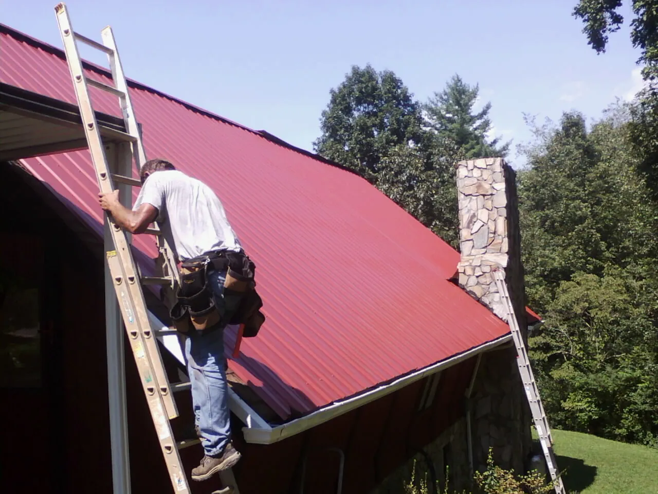 Roofer on a ladder beside a red metal roof and stone chimney — Soffit & Fascia Repair by Roofing Brothers in Winamac